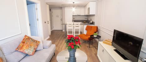 Een moderne woonkamer met een bank, een bijzettafeltje met een vaas, een televisie en een keuken op de achtergrond.