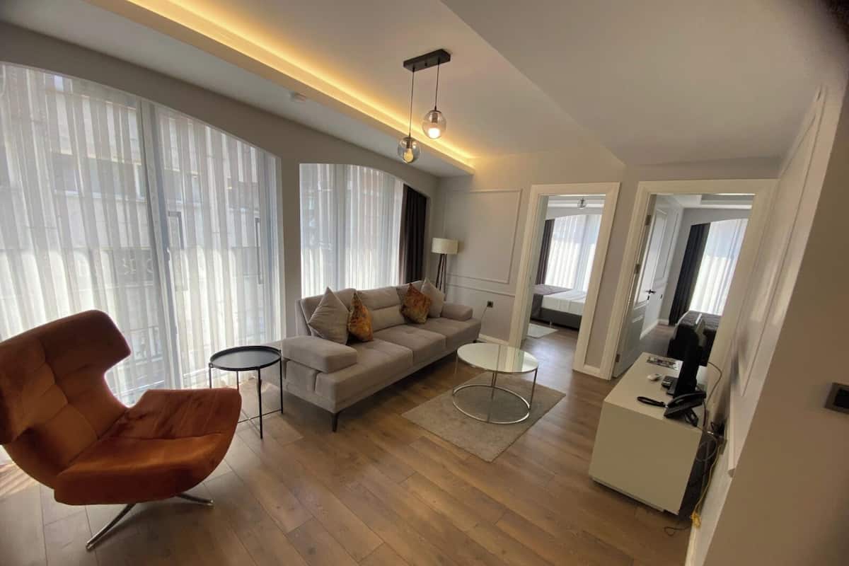 A modern living room with a sofa, a round coffee table, and a chair. There is a desk with a phone and a lamp. The room has large windows with blinds.