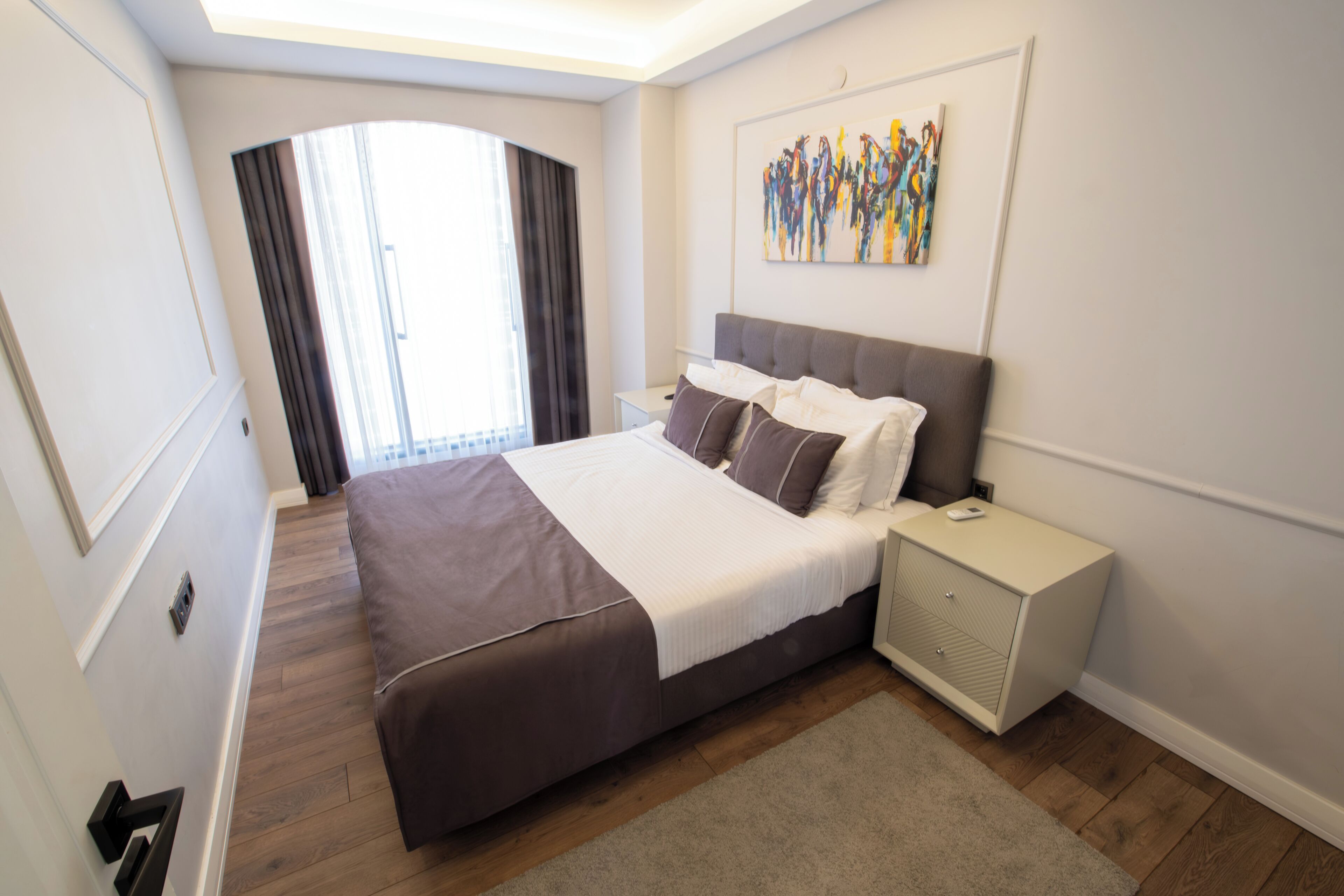 Una habitación de hotel moderna con una cama grande, una mesita de noche, cortinas en la ventana y un cuadro en la pared.