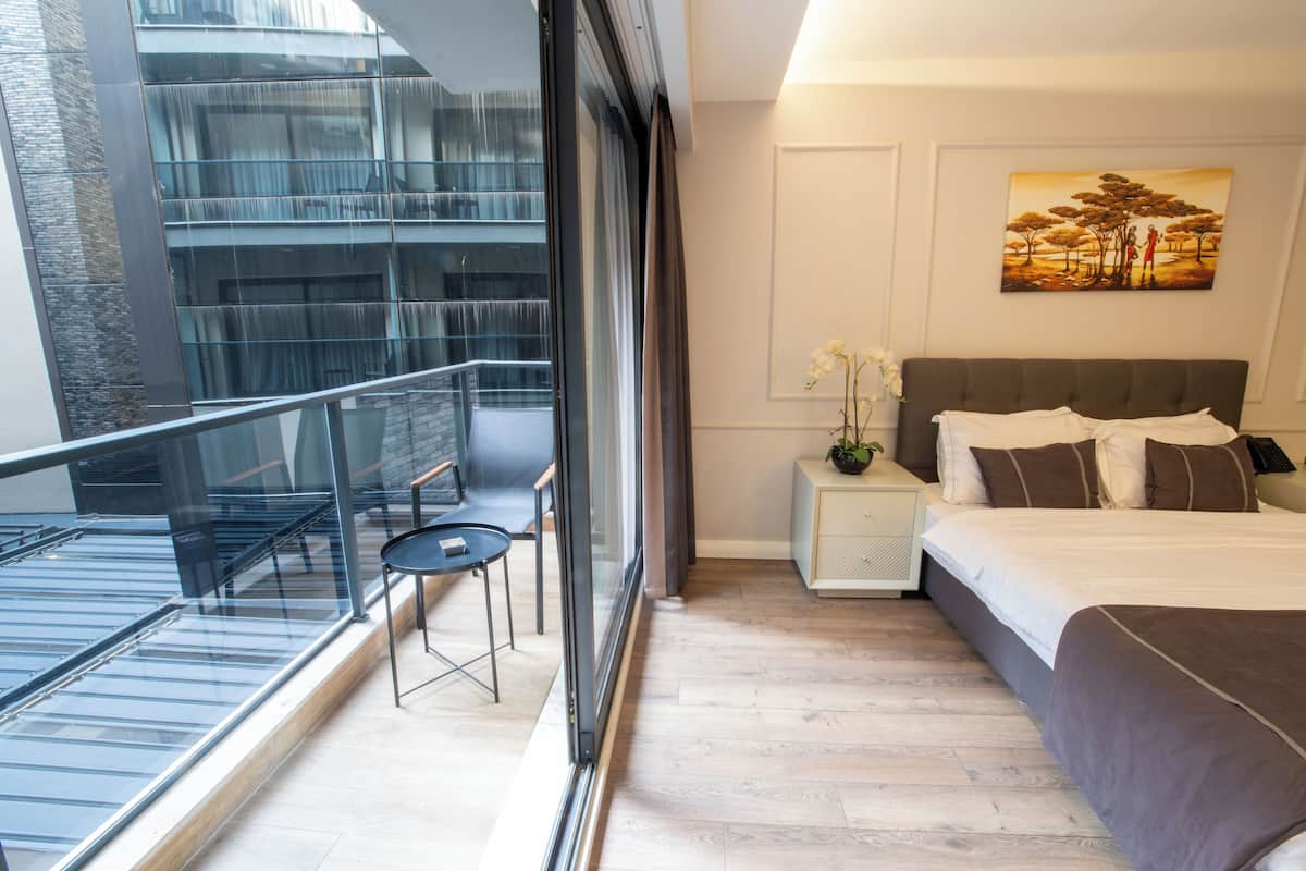 A modern hotel room with a large bed, a small table, a nightstand, a painting on the wall, and a balcony with a glass railing.