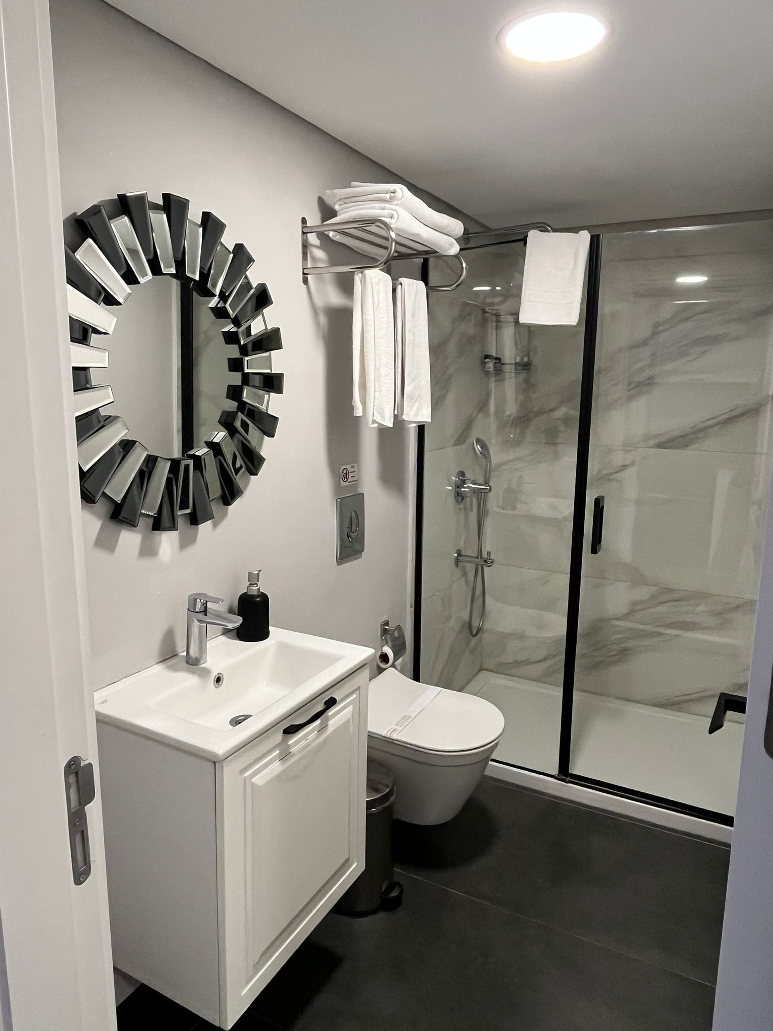 A modern bathroom with a white vanity, a round mirror, a glass shower enclosure, and a toilet.