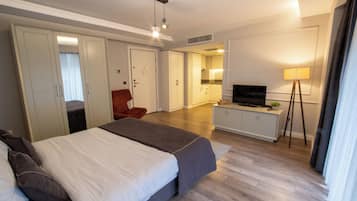 A modern hotel room with a large bed, a desk with a TV, a wardrobe, and a kitchenette.