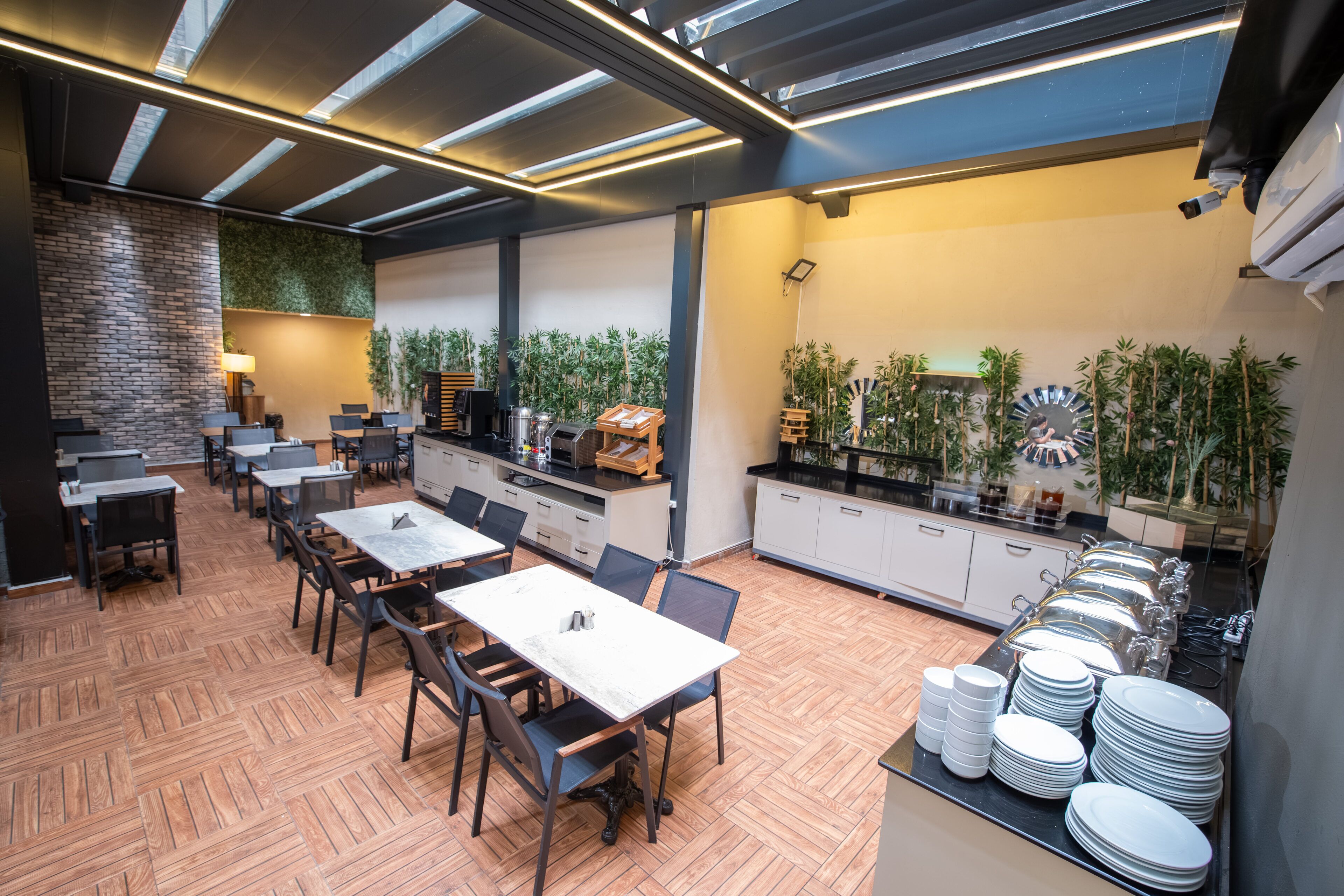 A modern dining area with wooden flooring, tables, chairs, and a kitchen area with plants and kitchenware.