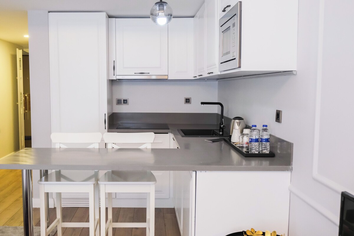A compact kitchen with white cabinetry, a stainless steel countertop, a kettle, and two bar stools.