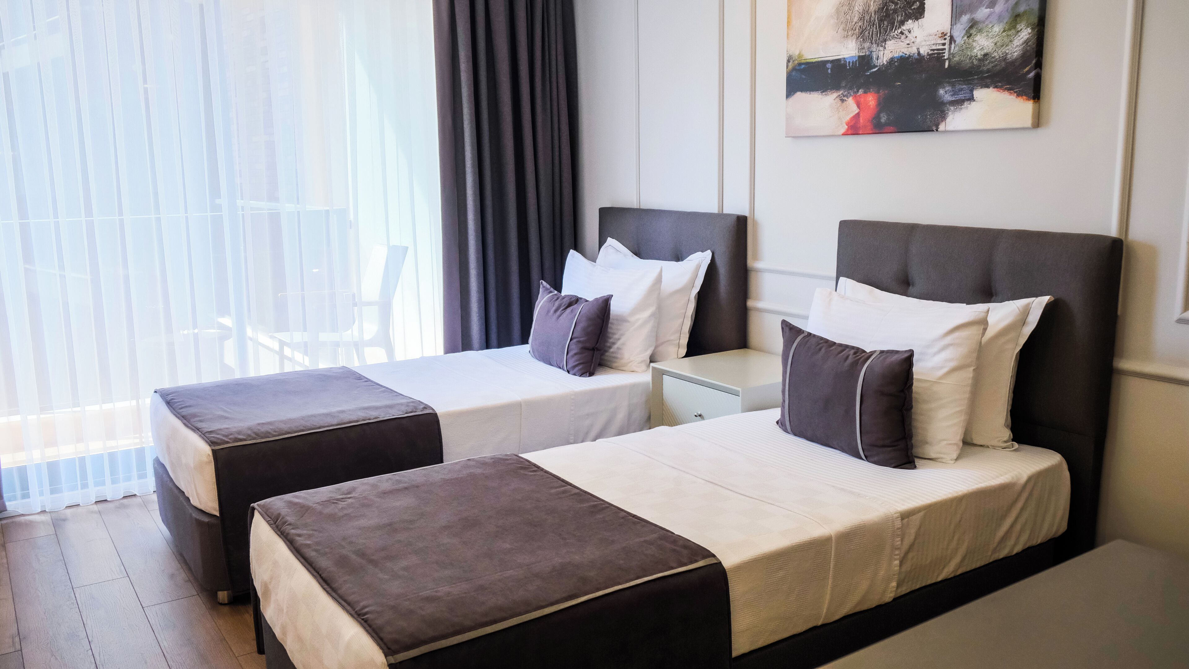 A hotel room with two beds, a nightstand, a painting on the wall, and a view of the outdoors through large windows.
