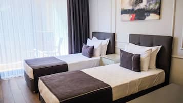 A hotel room with two beds, a nightstand, a painting on the wall, and a view of the outdoors through large windows.