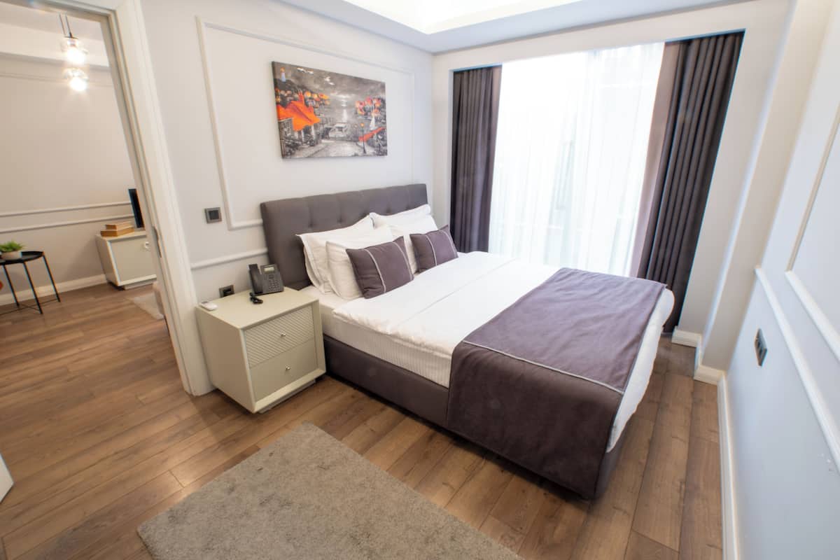 A modern bedroom with a large bed, a bedside table, a nightstand, a window with curtains, and a painting on the wall.