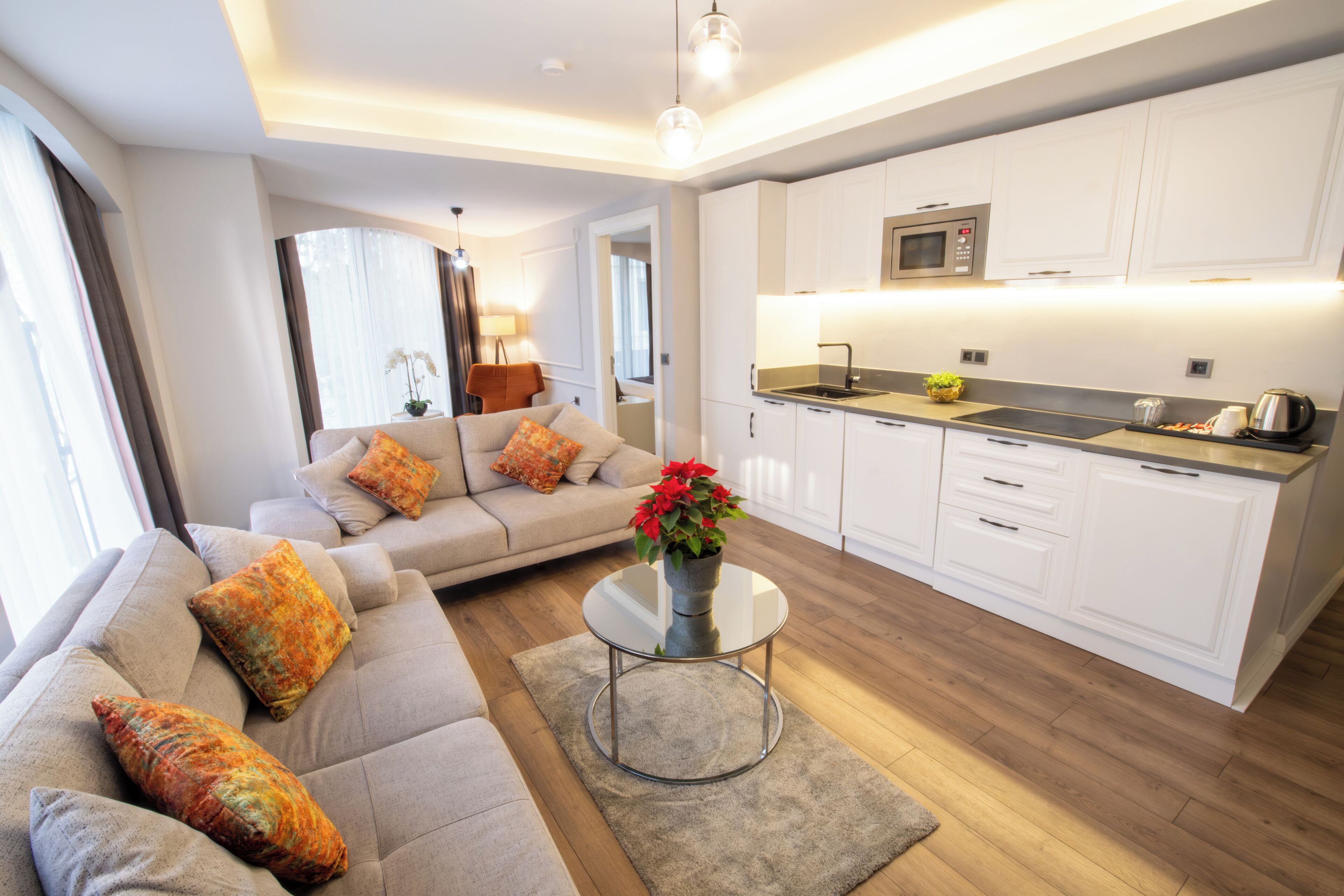 A modern living room with a sofa, a coffee table with a vase of flowers, and a kitchen area with white cabinets and a microwave.