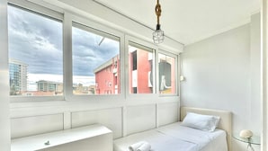 Une chambre avec un lit simple, un banc blanc et une table en verre agrémentée d’un objet décoratif.