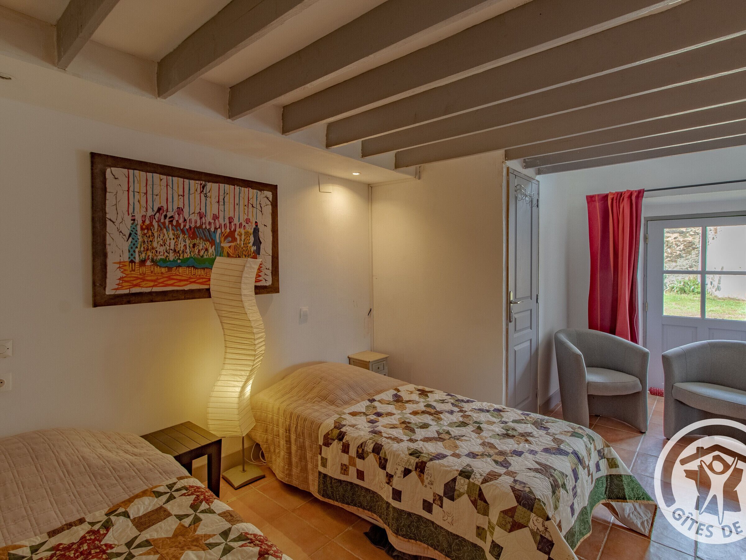 A bedroom with two beds, a wall-mounted painting, a standing lamp, and a door leading to a balcony with a red curtain.