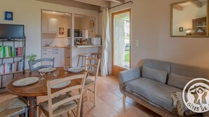 Una sala de estar acogedora con un sofá gris, una mesa de comedor de madera con sillas, una estantería y una cocina visible a través de una puerta abierta.