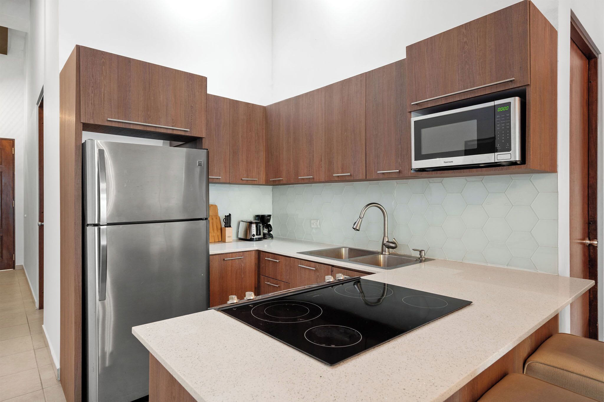 A modern kitchen with wooden cabinets, a stainless steel refrigerator, a microwave, a sink, and a black induction cooktop.