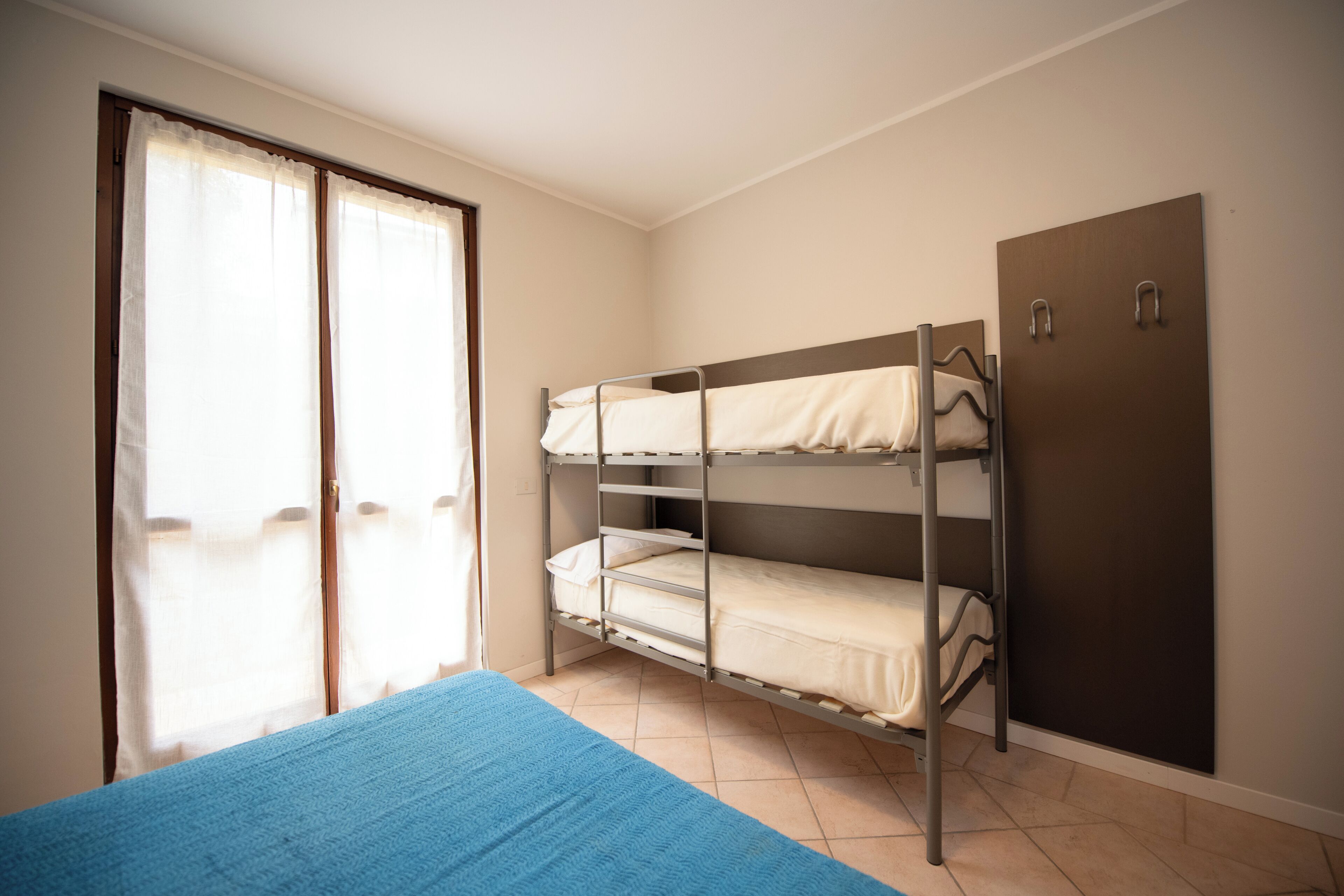 A bunk bed room with a blue bedspread, a wardrobe, and a window with curtains.