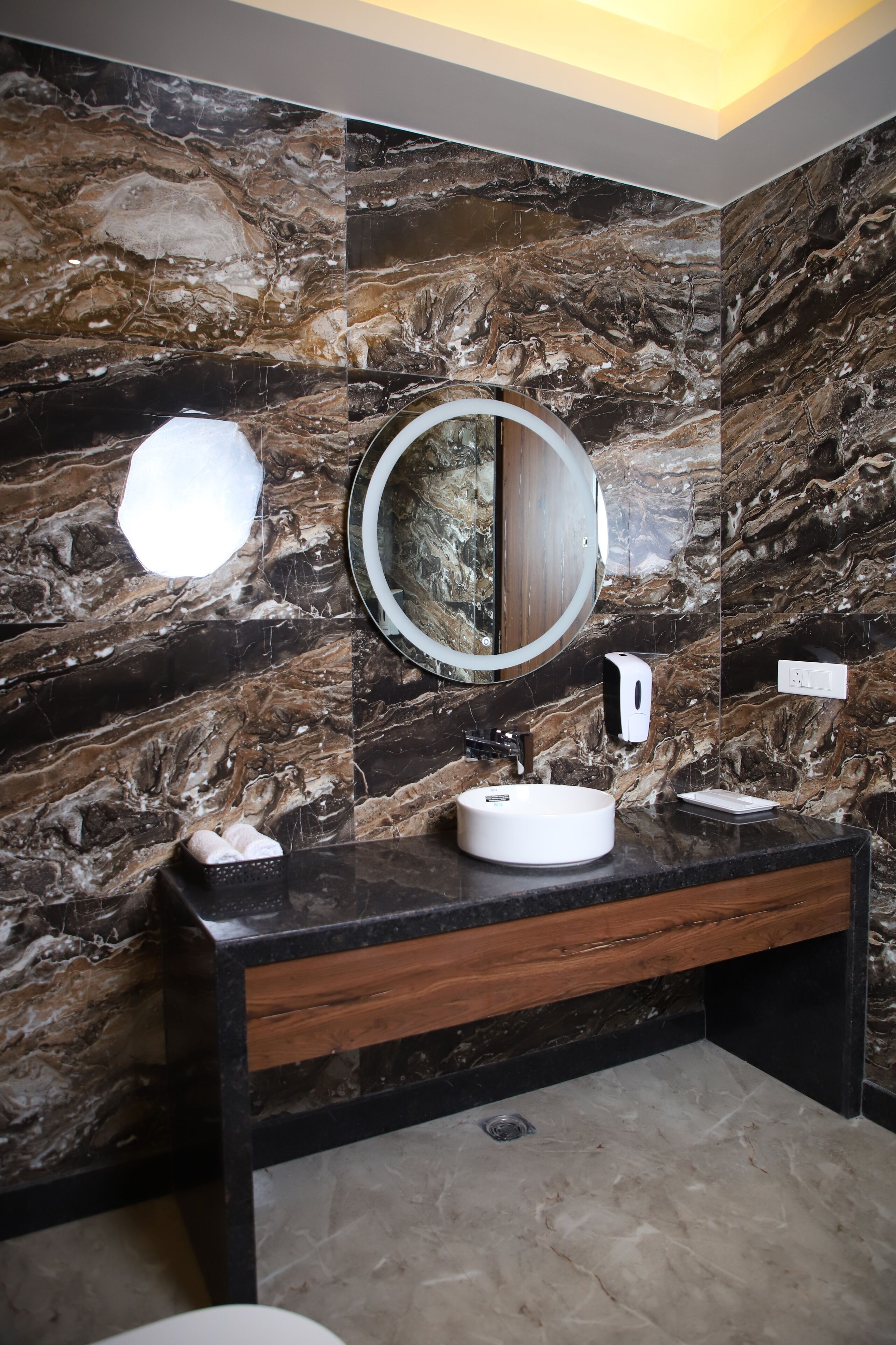 Une salle de bain avec un mur en marbre, un miroir rond, un lavabo blanc et un distributeur de savon.