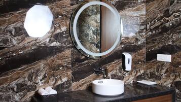 Une salle de bain avec un mur en marbre, un miroir rond, un lavabo blanc et un distributeur de savon.