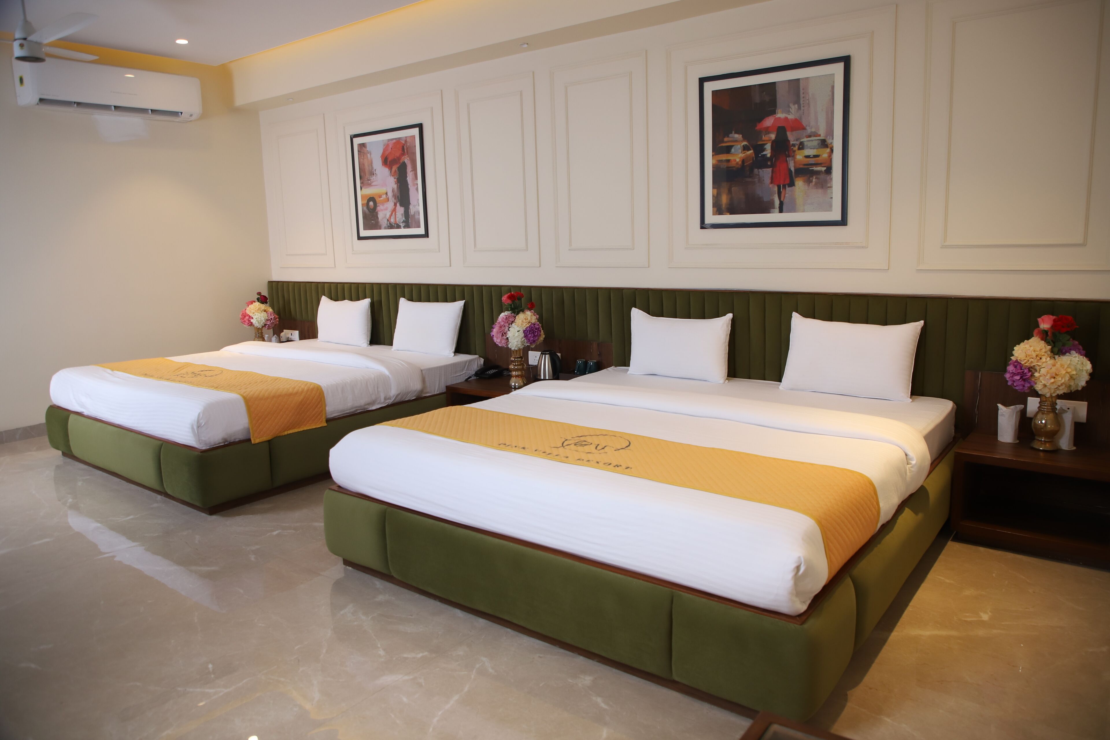 Habitación de hotel con dos camas, cada una con cabeceros verdes y ropa de cama blanca. En la pared hay cuadros enmarcados, una mesita de noche con un jarrón de flores y un ventilador de techo.