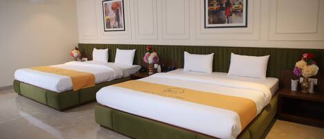 Habitación de hotel con dos camas, cada una con cabeceros verdes y ropa de cama blanca. En la pared hay cuadros enmarcados, una mesita de noche con un jarrón de flores y un ventilador de techo.