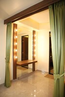 A room with a vanity mirror surrounded by lights, a wooden bench, and green curtains.