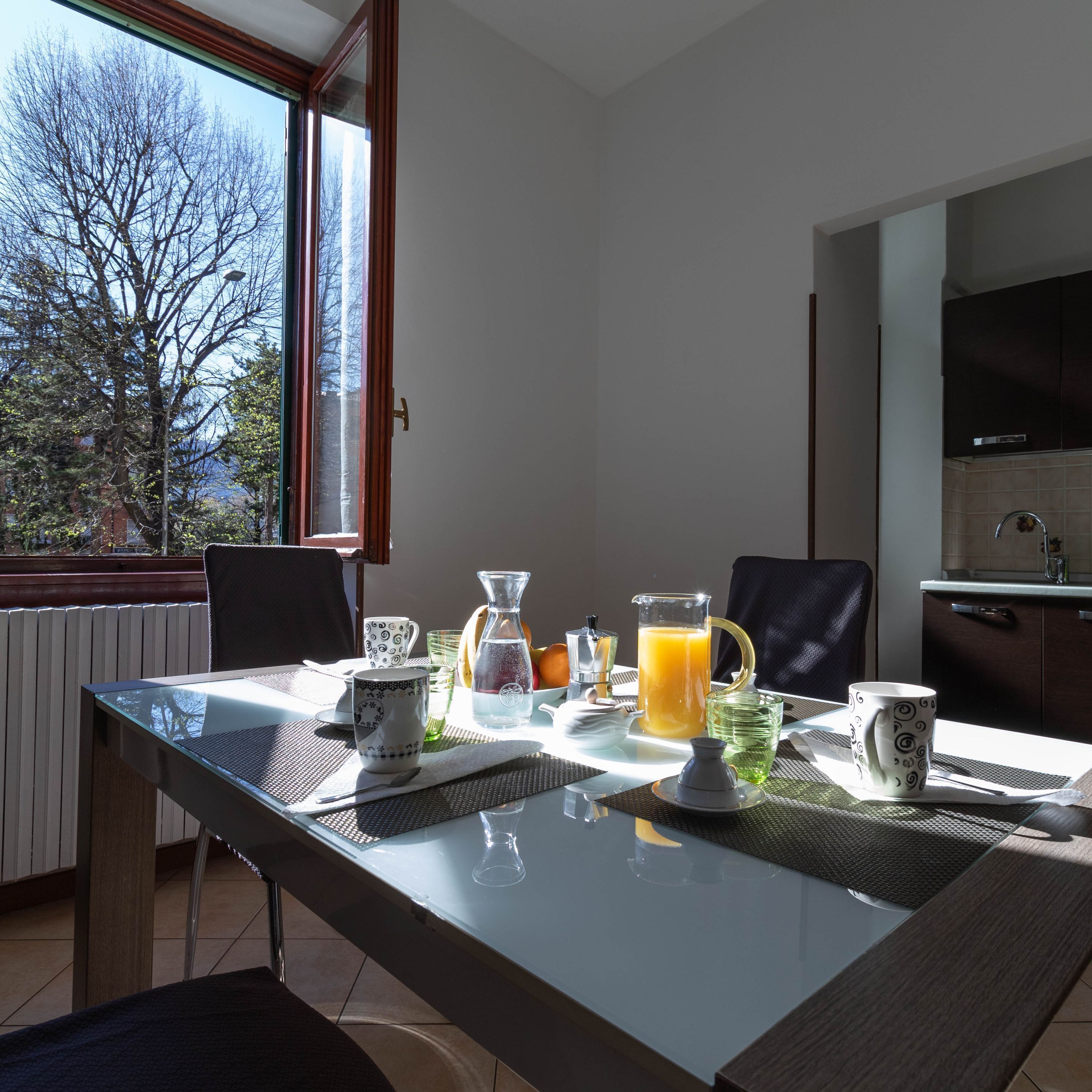 A breakfast table with a pitcher of orange juice, a coffee pot, and a mug, set on a balcony with a view of trees and a building.