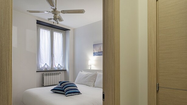 A bedroom with a bed, a ceiling fan, a window with curtains, a radiator, and a painting on the wall. - Comfy Apartment Near the Harbor by Wonderful Italy (Genoa)
