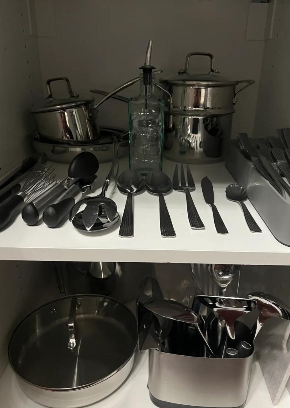 A kitchen with stainless steel pots, a glass bottle, and utensils neatly arranged on a white shelf.
