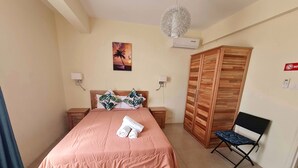 A neatly arranged bedroom with a bed, a wooden wardrobe, a folding chair, and a painting on the wall. - Le Mirage Cozy Beach Apartment, 5 min walk to the beach. Enjoy sunset view (Flic en Flac)