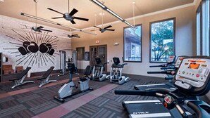 Un gimnasio con diversos equipos de ejercicio, ventiladores de techo y amplias ventanas.