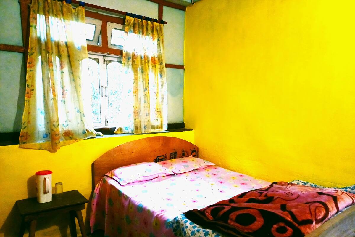 A small room with a bed, a window with floral curtains, a small table with a cup and a candle, and a yellow wall.