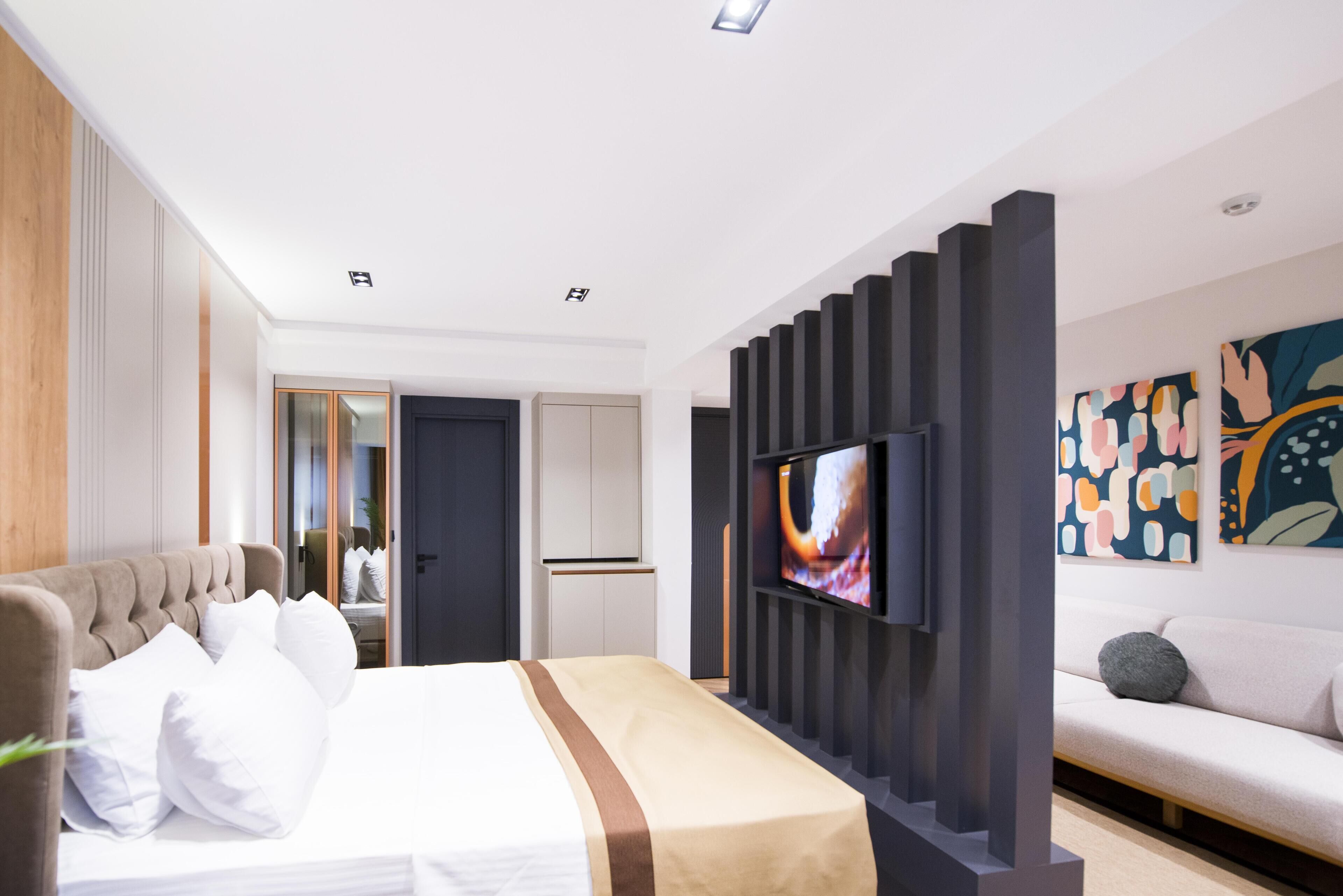 Une chambre d’hôtel moderne avec un grand lit, un téléviseur à écran plat fixé sur un panneau sombre, un canapé et des œuvres d’art abstraites accrochées au mur.