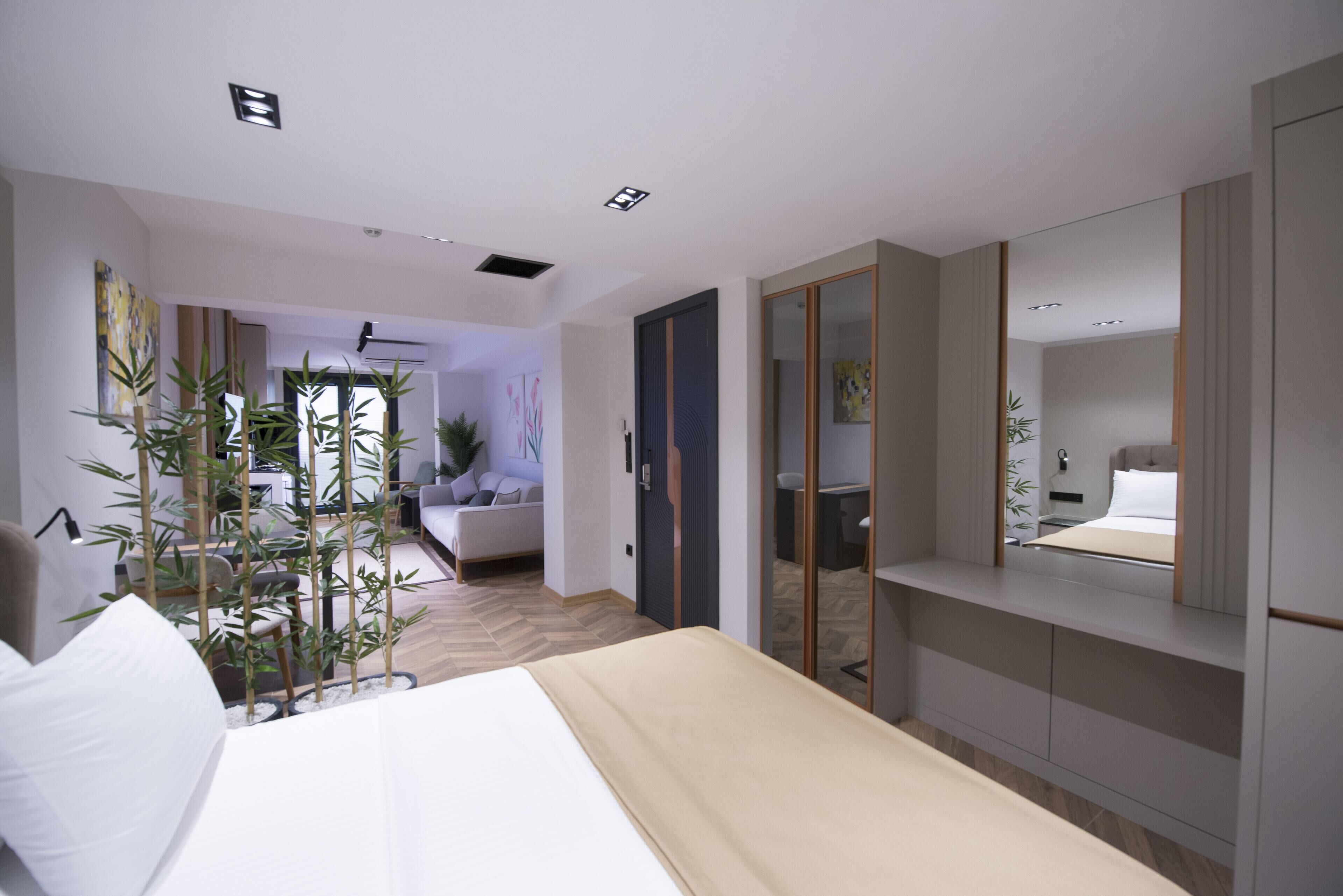 Una habitación de hotel moderna con cama, mesita de noche y un espejo grande. Hay una planta en maceta y vistas del área de estar.