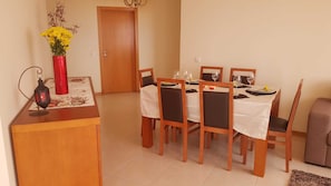 Een eetruimte met een houten tafel voor vier personen, omringd door stoelen. Op de tafel staat een rode vaas met gele bloemen, en op het buffet een kleine lantaarn.