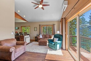 Un salon spacieux doté d’un ventilateur de plafond, d’une cheminée et d’une grande fenêtre donnant sur la forêt.