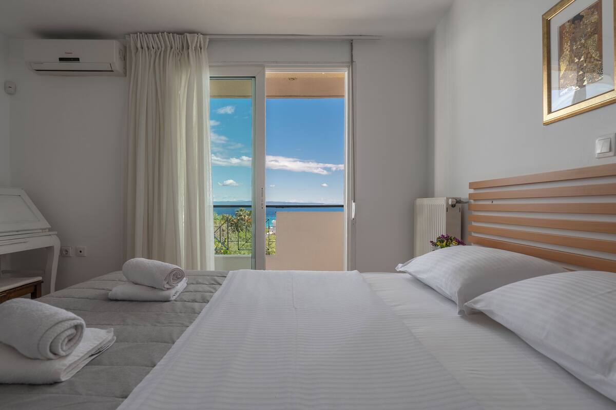 A hotel room with a bed, a desk, a chair, a window with curtains, and a view of the sea.