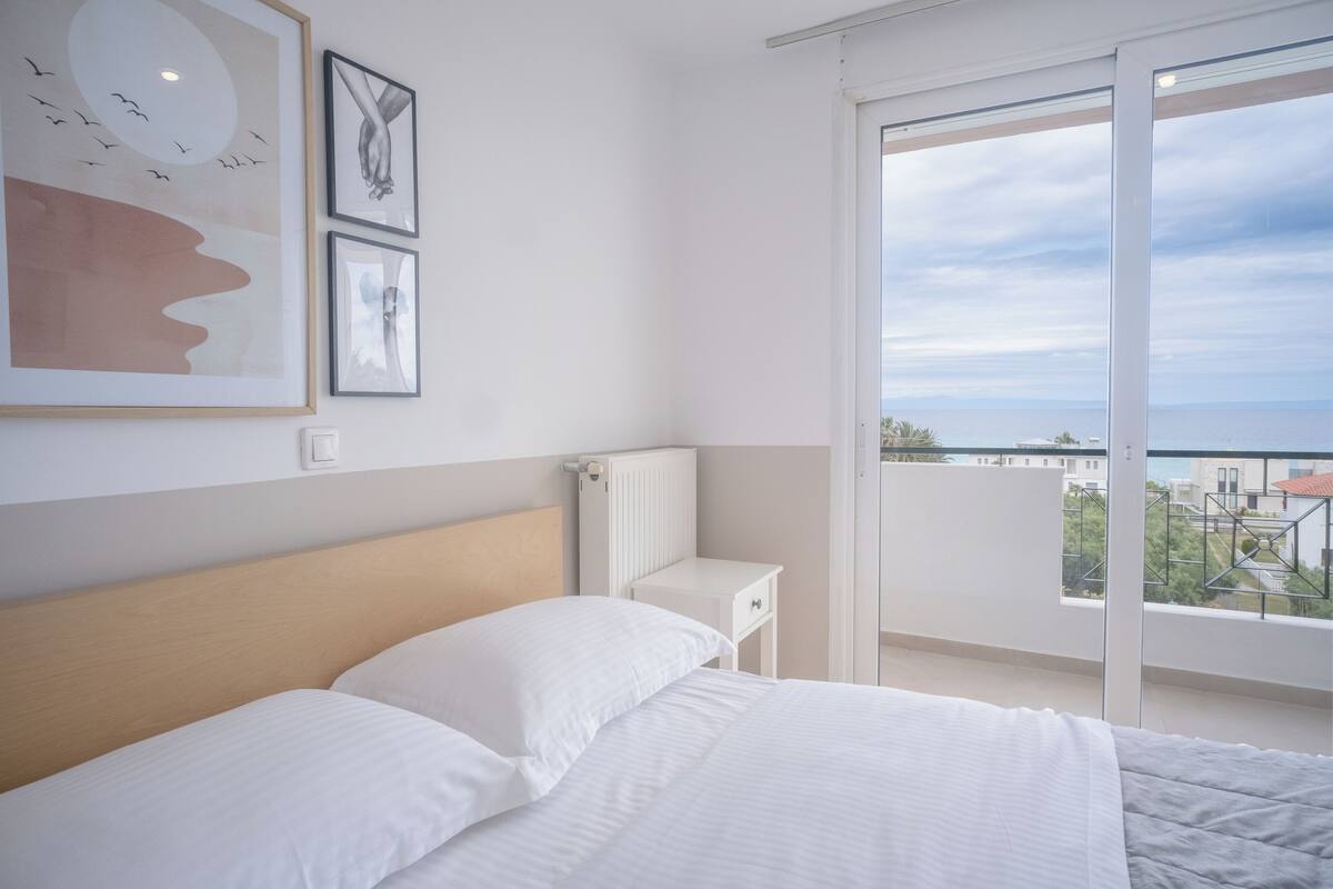 A bedroom with a large bed, a bedside table, and a view of the city through a balcony.