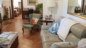 Een ruime woonkamer met een leren bank, fauteuils en een salontafel. Aan de muur hangt een grote spiegel en aan het plafond een kroonluchter.
