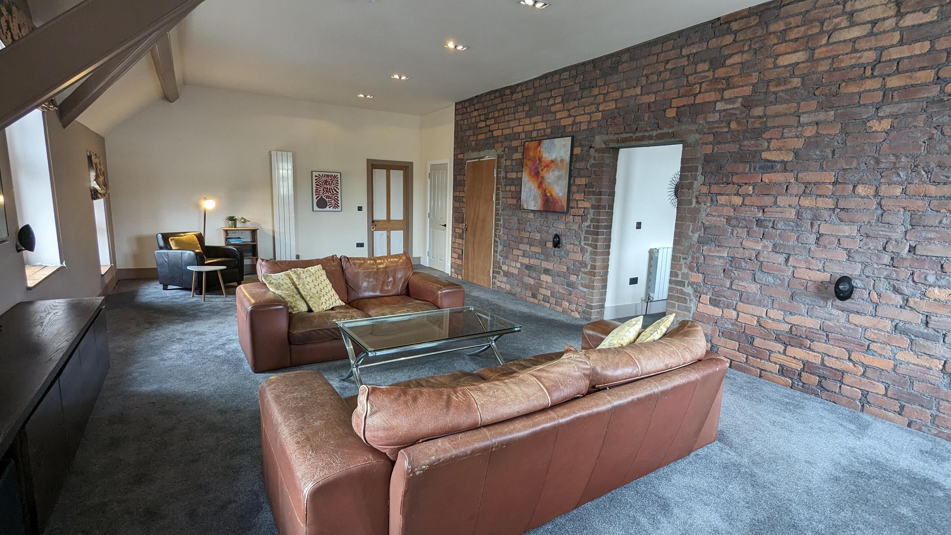 A spacious living room with a leather sofa, a glass coffee table, and a brick wall.