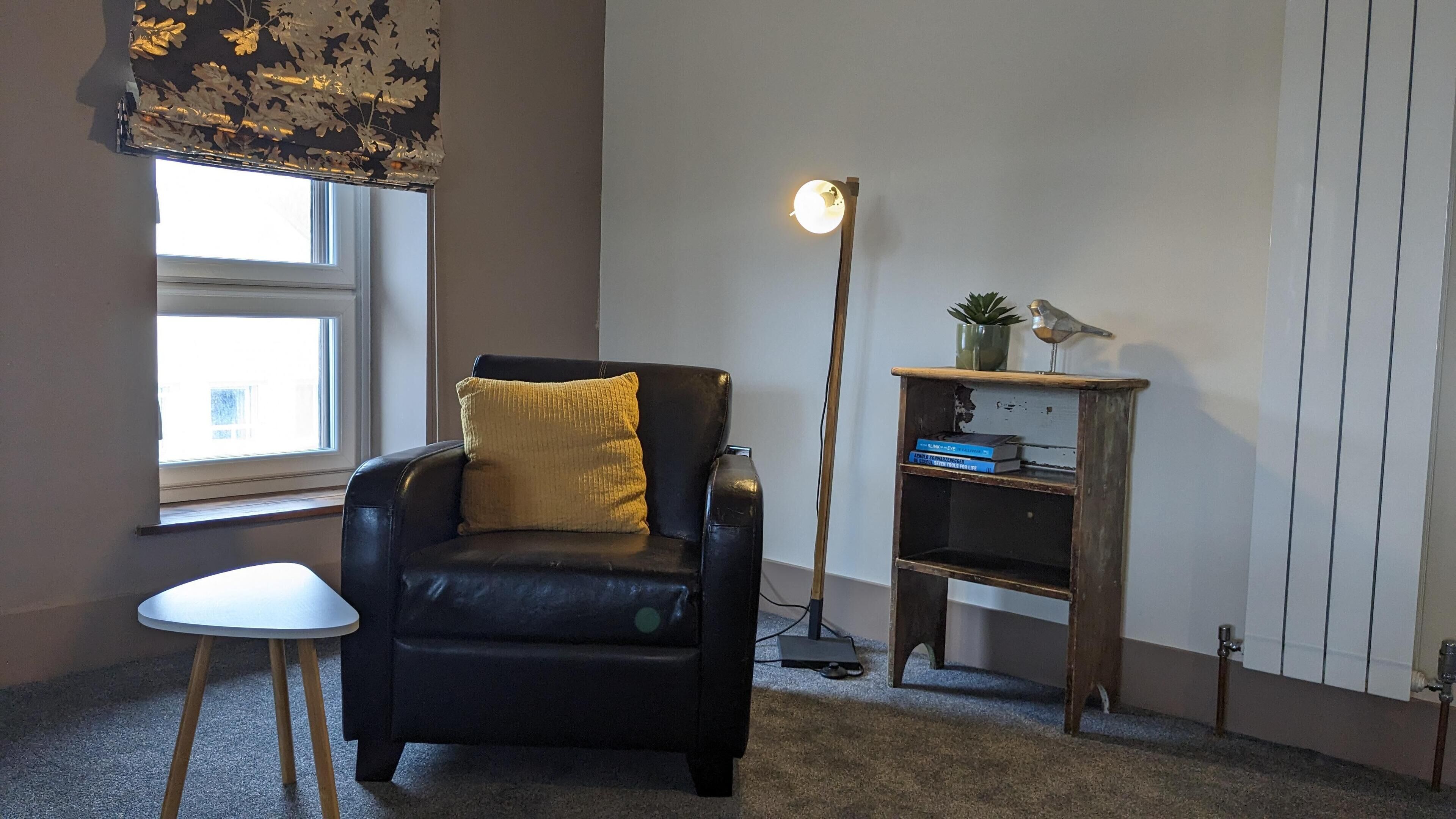 Une pièce comprenant un fauteuil en cuir noir, une petite table blanche, un lampadaire et une étagère en bois avec des livres et une plante.