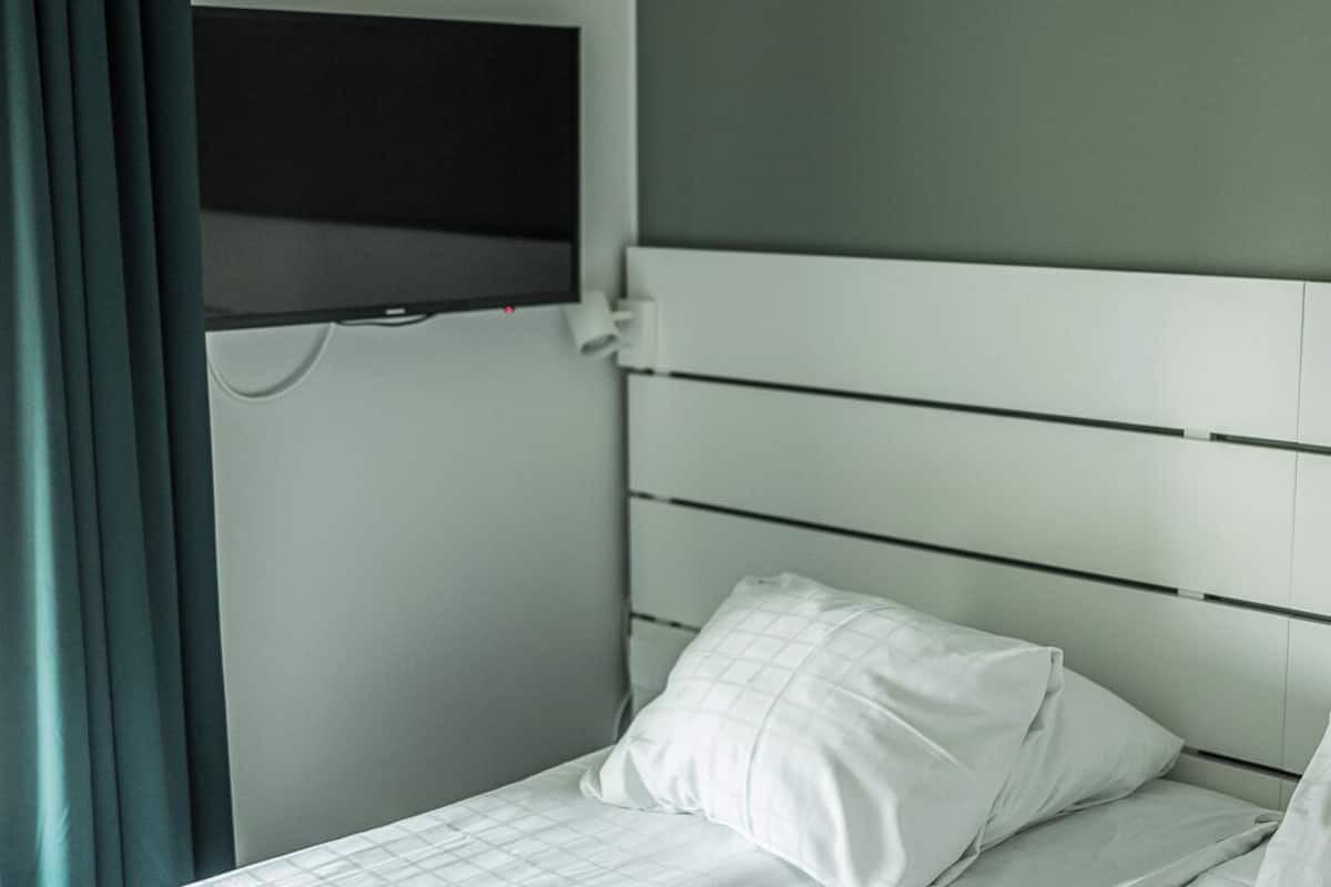 A single bed with a white headboard and a pillow, a flat-screen TV mounted on the wall, and a window with curtains.