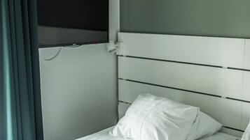 Una cama individual con cabecera blanca y almohada, un televisor de pantalla plana en la pared, y una ventana con cortinas.