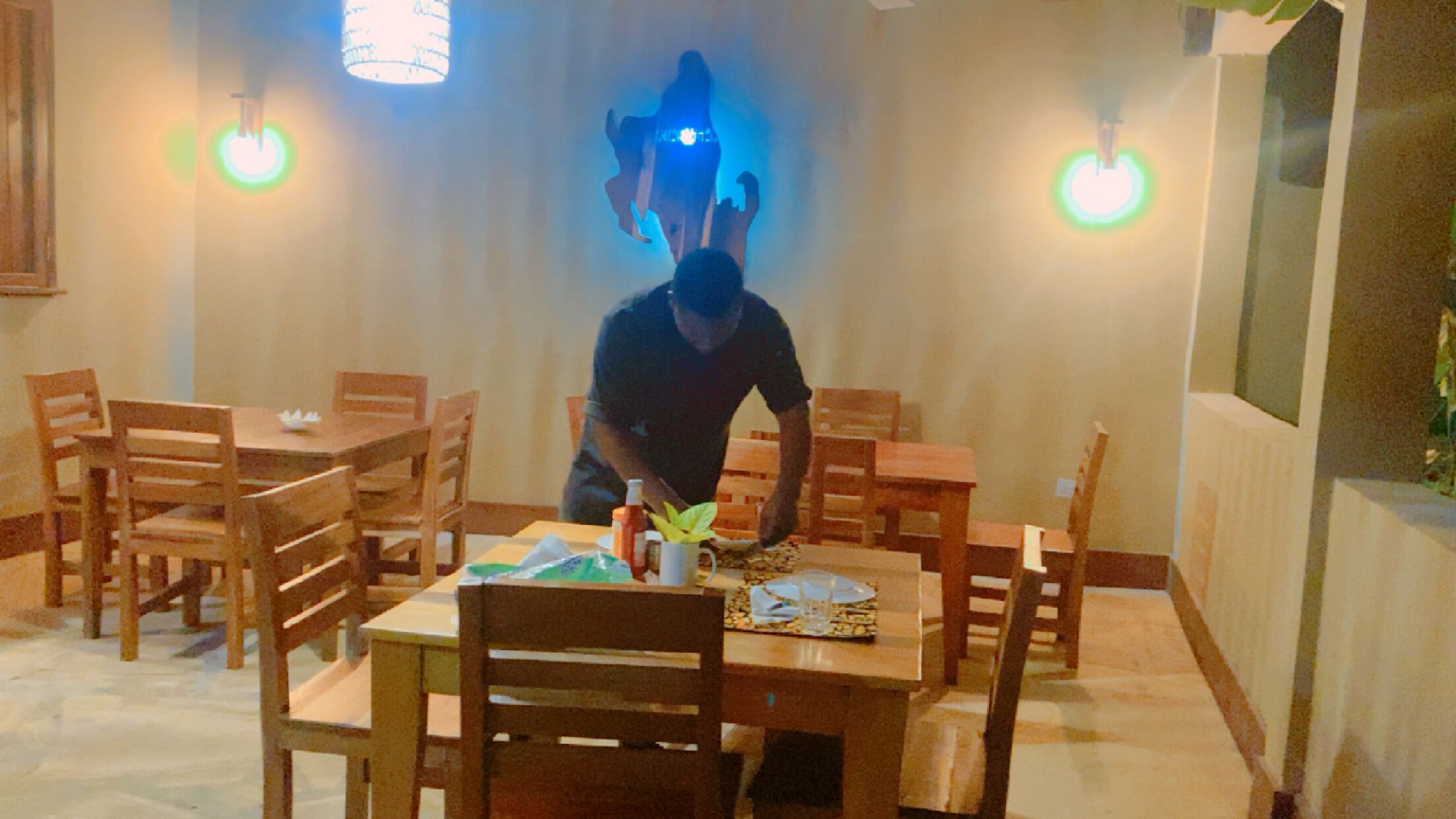 A dining area with wooden tables and chairs, a person serving food, and wall-mounted lights.