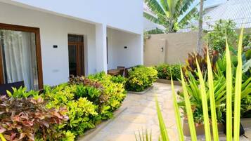 Um edifício moderno com fachada branca, um caminho de pedra e vegetação exuberante, incluindo bananeiras e arbustos diversos.