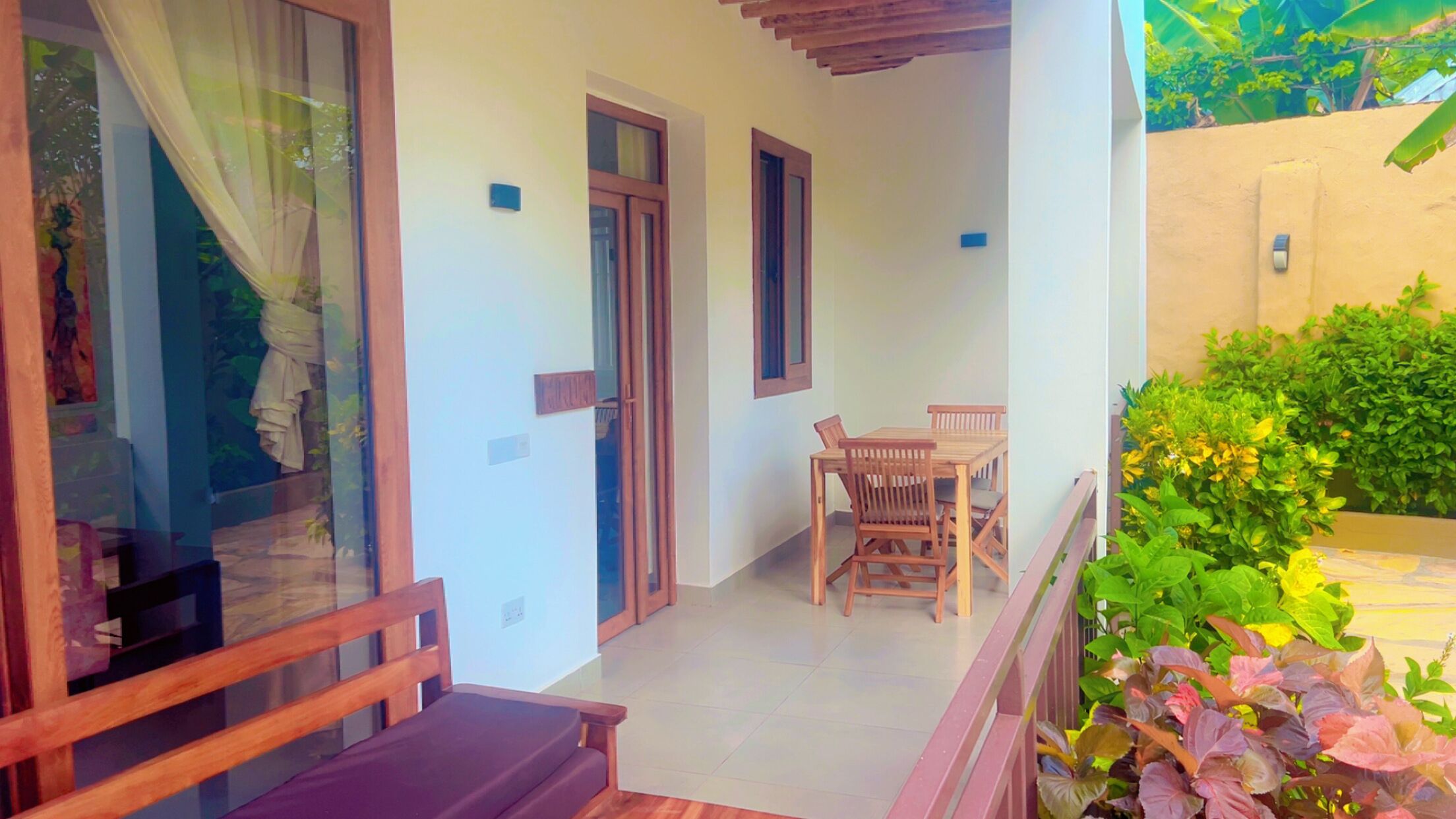 A balcony with a wooden table and chairs, a glass door, and a view of greenery.