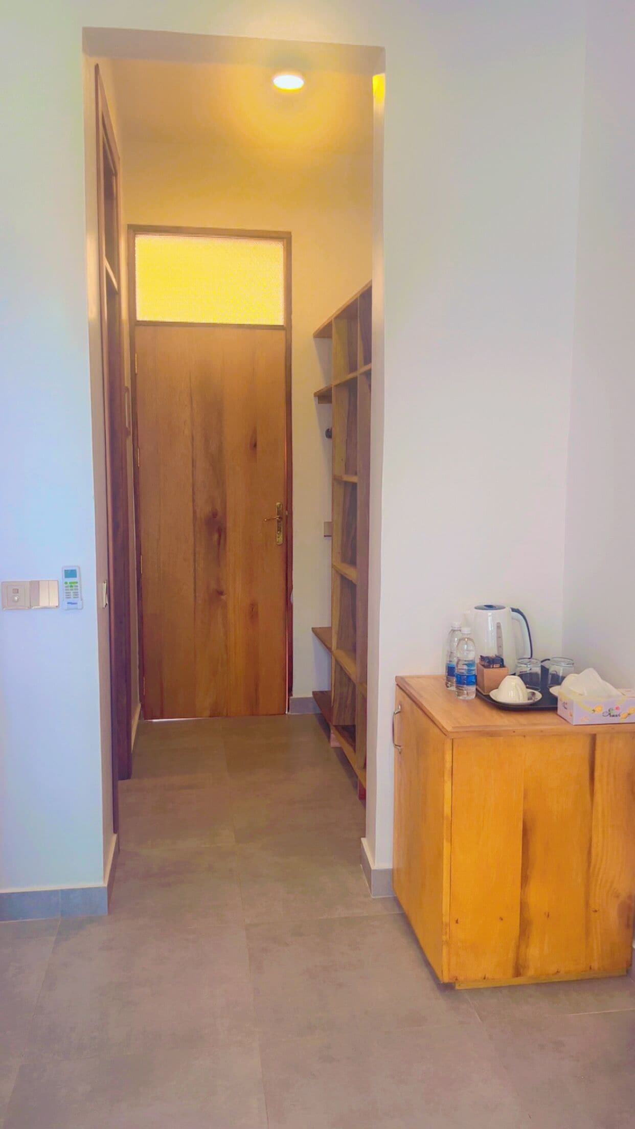 Un pasillo con una puerta de madera al final, una pequeña cocina americana con una cafetera, y una estantería con diversos objetos.