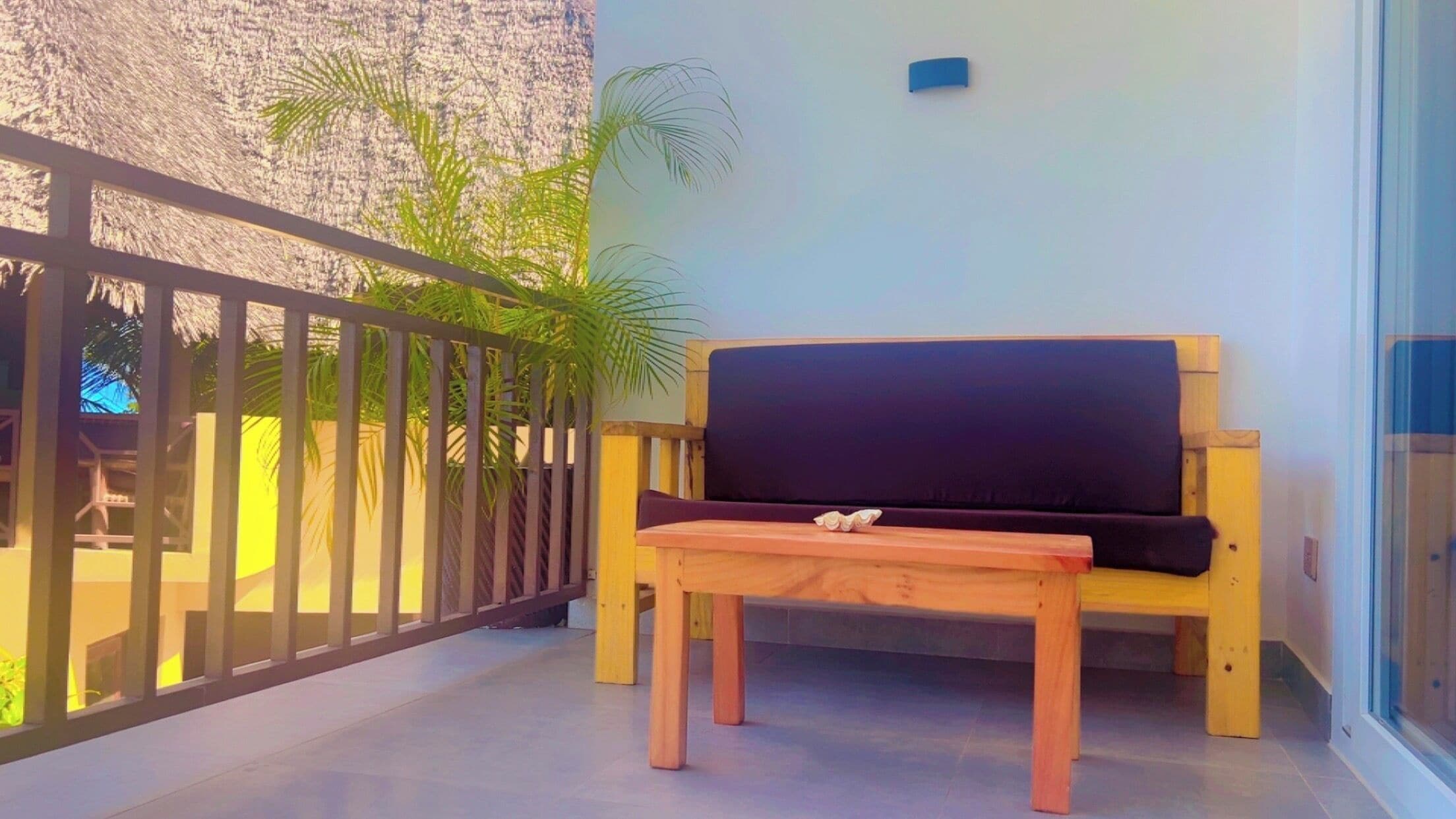 A balcony with a wooden bench and a small table, a railing, and a palm tree.