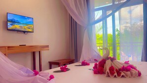 A hotel room with a bed, a TV on the wall, and a view of greenery outside. - The Dream Village (Kiwengwa)