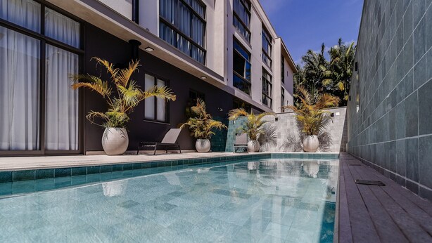 Área junto à piscina, com piscina, plantas em vasos e um edifício com janelas amplas.