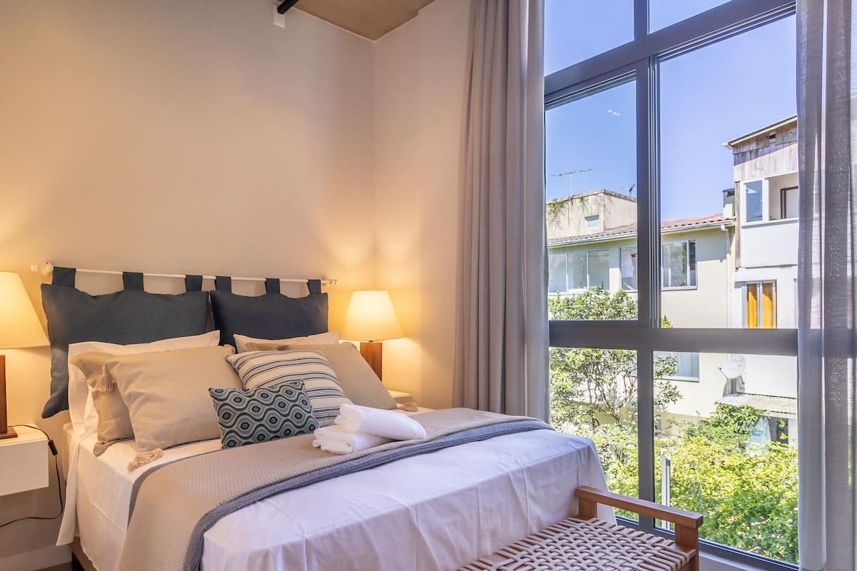 A hotel room with a bed, two bedside lamps, a window with a view of buildings, and a chair by the window.