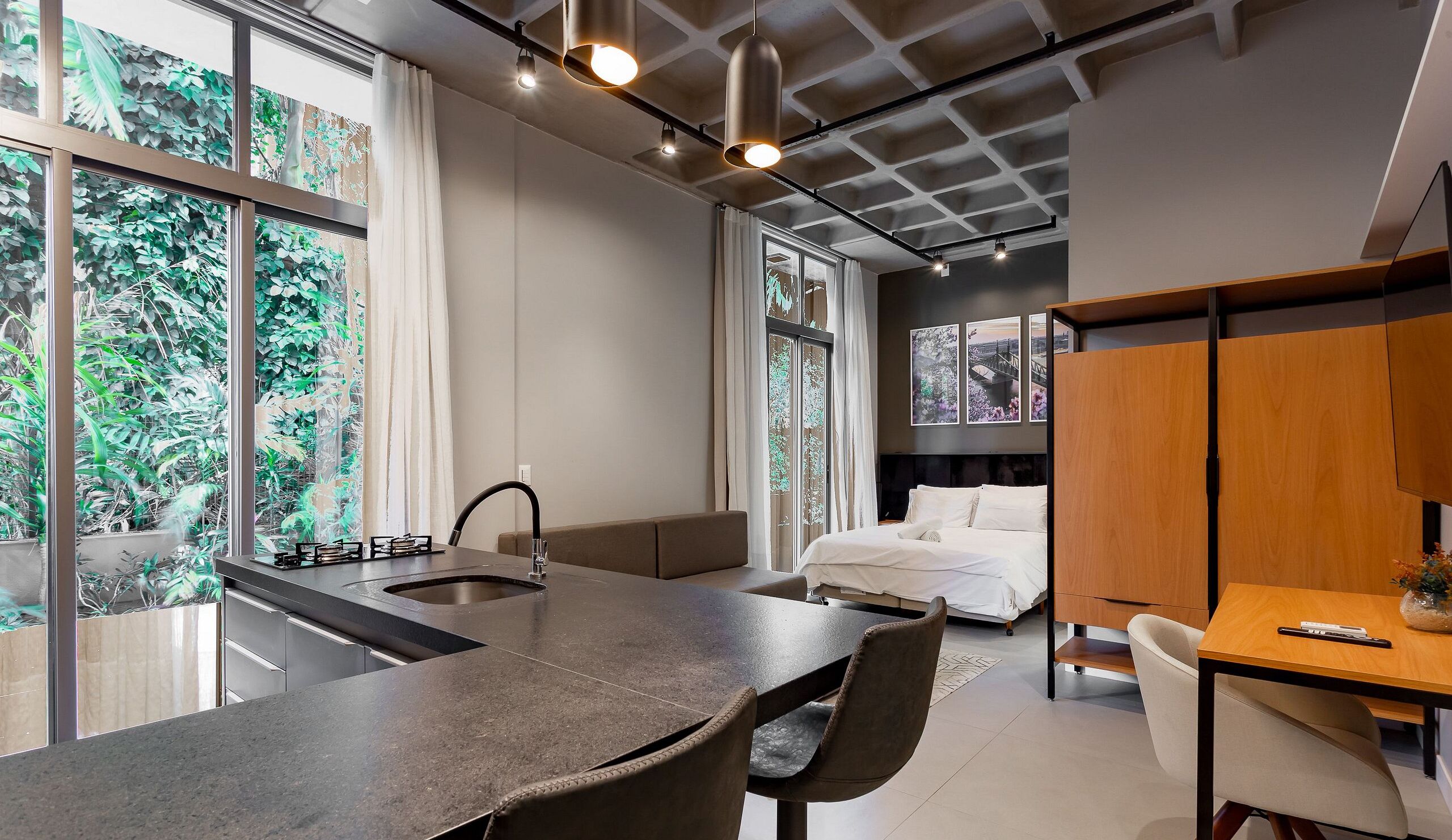 Una cocina moderna con una isla central, un área de comedor con mesa y sillas, una cama con cabecera, y ventanas amplias con vista a zonas verdes.