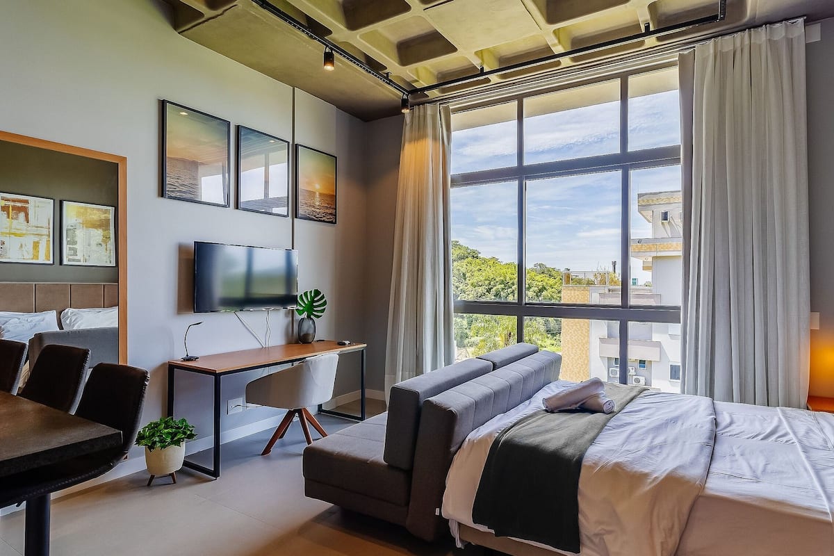 A modern hotel room with a bed, sofa, desk, TV, and large windows.