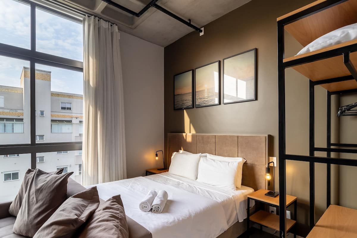 A hotel room with a bed, a desk, a chair, a bedside table, a lamp, and a view of buildings outside.